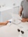 A person is making a payment using a credit card on a small, square card reader connected to a modern point-of-sale system at a minimalist counter. Another person is standing nearby, possibly a cashier, wearing a grey t-shirt. Sunglasses rest on the counter, and the background features a simple, white decor.