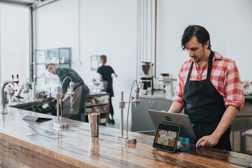 A man in a red checkered shirt and black apron is standing at a wooden countertop using a tablet device. In the background, a modern coffee shop setting is visible with another person working at a counter and various coffee-making equipment around.