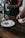 A person is using a small card reader to process a payment in a cafe setting. On the wooden table, there is a white plate with a glass of milk and a chocolate chip cookie. Another person is holding a glass jar under a water tap.