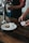 A person is using a small card reader to process a payment in a cafe setting. On the wooden table, there is a white plate with a glass of milk and a chocolate chip cookie. Another person is holding a glass jar under a water tap.