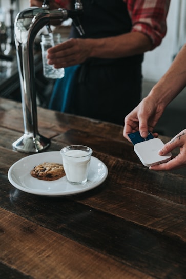 A person is using a small card reader to process a payment in a cafe setting. On the wooden table, there is a white plate with a glass of milk and a chocolate chip cookie. Another person is holding a glass jar under a water tap.