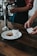 A person is using a small card reader to process a payment in a cafe setting. On the wooden table, there is a white plate with a glass of milk and a chocolate chip cookie. Another person is holding a glass jar under a water tap.
