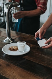 A person is using a small card reader to process a payment in a cafe setting. On the wooden table, there is a white plate with a glass of milk and a chocolate chip cookie. Another person is holding a glass jar under a water tap.