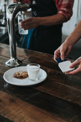 A person is using a small card reader to process a payment in a cafe setting. On the wooden table, there is a white plate with a glass of milk and a chocolate chip cookie. Another person is holding a glass jar under a water tap.