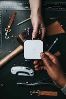 A person processes a payment using a contactless card reader while another hand holds a credit card. The scene is set on a dark surface with various leather crafting tools and materials scattered around, including a ruler, leather straps, and a utility knife.