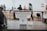 A modern point-of-sale system is positioned on a counter in a contemporary café or work environment, with a few markers on the left. The background features people engaging and moving around, giving a sense of activity and interaction.