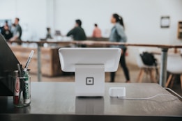 A modern point-of-sale system is positioned on a counter in a contemporary caf&eacute; or work environment, with a few markers on the left. The background features people engaging and moving around, giving a sense of activity and interaction.