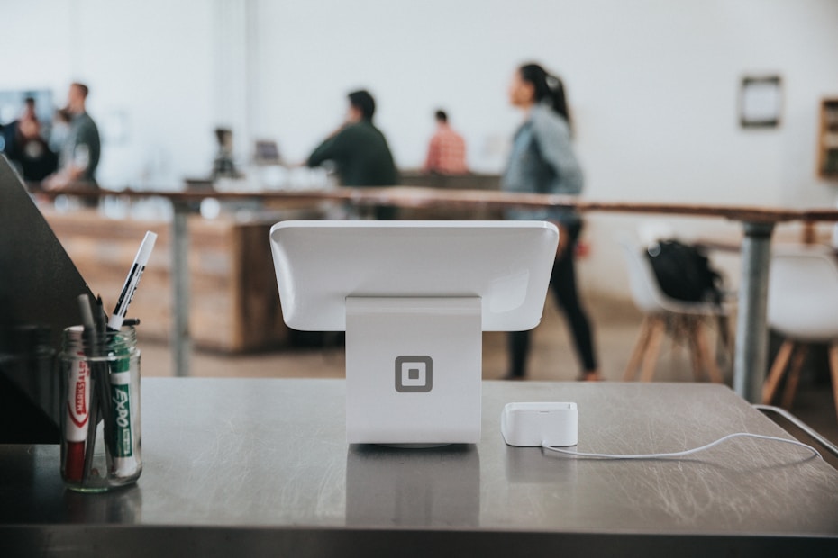 A modern point-of-sale system is positioned on a counter in a contemporary café or work environment, with a few markers on the left. The background features people engaging and moving around, giving a sense of activity and interaction.