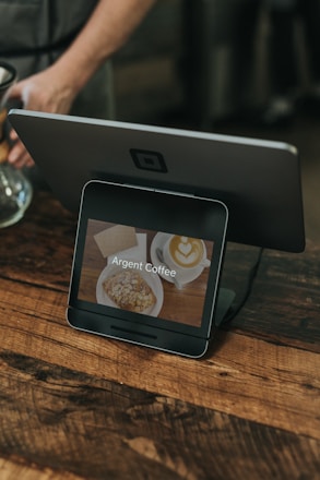 A digital point-of-sale device is displayed on a wooden counter. The screen shows an image of a latte with heart-shaped latte art and a pastry, accompanied by the text 'Argent Coffee.' Part of a person's arm is visible in the background, suggesting a caf&eacute; setting.