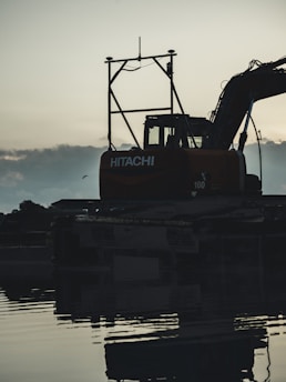 An excavator silhouetted against the sky, positioned near a body of water with its reflection visible. The machine is clearly marked with branding and is set in a calm and serene environment, as the sun appears to set or rise in the background, casting soft light and shadows.
