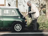 An elderly man wearing a beige jacket and glasses is inspecting the rear of a vintage green Mini Cooper parked on a roadside. The setting is outdoors with greenery and trees in the background, suggesting a serene, countryside environment.