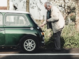 An elderly man wearing a beige jacket and glasses is inspecting the rear of a vintage green Mini Cooper parked on a roadside. The setting is outdoors with greenery and trees in the background, suggesting a serene, countryside environment.