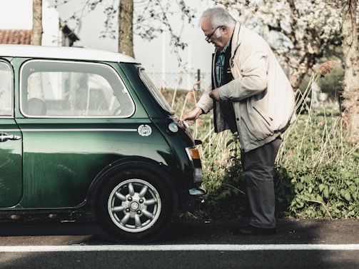 An elderly man wearing a beige jacket and glasses is inspecting the rear of a vintage green Mini Cooper parked on a roadside. The setting is outdoors with greenery and trees in the background, suggesting a serene, countryside environment.