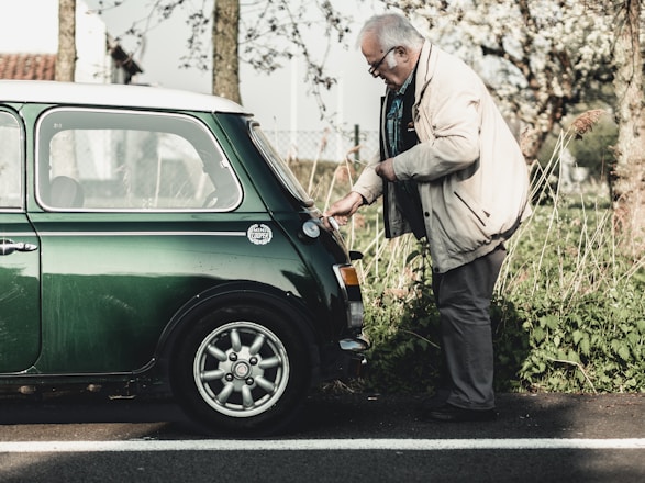 An elderly man wearing a beige jacket and glasses is inspecting the rear of a vintage green Mini Cooper parked on a roadside. The setting is outdoors with greenery and trees in the background, suggesting a serene, countryside environment.