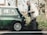An elderly man wearing a beige jacket and glasses is inspecting the rear of a vintage green Mini Cooper parked on a roadside. The setting is outdoors with greenery and trees in the background, suggesting a serene, countryside environment.