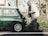 An elderly man wearing a beige jacket and glasses is inspecting the rear of a vintage green Mini Cooper parked on a roadside. The setting is outdoors with greenery and trees in the background, suggesting a serene, countryside environment.