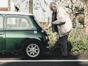 An elderly man wearing a beige jacket and glasses is inspecting the rear of a vintage green Mini Cooper parked on a roadside. The setting is outdoors with greenery and trees in the background, suggesting a serene, countryside environment.