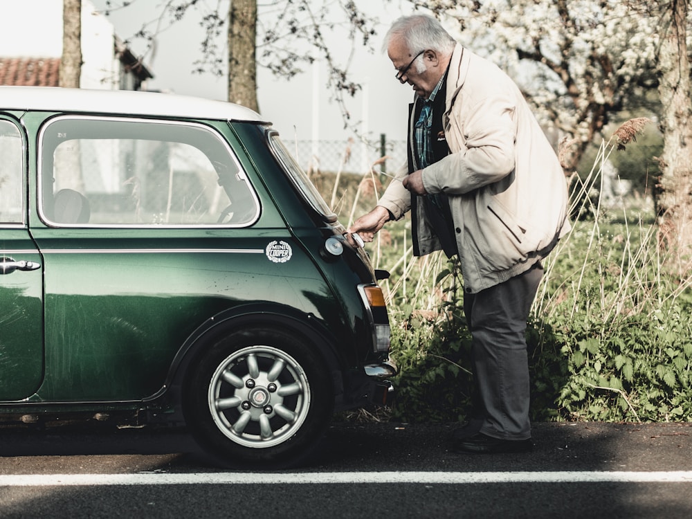 An elderly man wearing a beige jacket and glasses is inspecting the rear of a vintage green Mini Cooper parked on a roadside. The setting is outdoors with greenery and trees in the background, suggesting a serene, countryside environment.