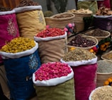 Colorful fabric bags and woven baskets display a variety of dried flowers and spices. The containers are filled with different colors and textures, showcasing ingredients like dried petals and herbs. Each bag is decorated with floral embroidery, enhancing the aesthetic presentation.