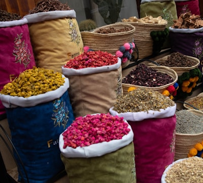 Colorful fabric bags and woven baskets display a variety of dried flowers and spices. The containers are filled with different colors and textures, showcasing ingredients like dried petals and herbs. Each bag is decorated with floral embroidery, enhancing the aesthetic presentation.