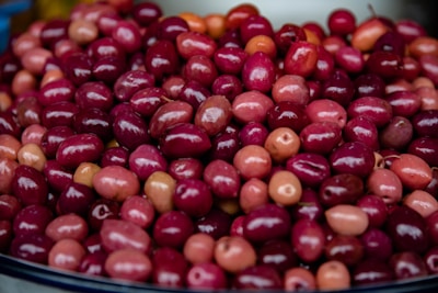 A pile of vibrant, glossy olives of varying shades of red and pink is displayed. The fruits are closely packed together, showcasing a rich, shiny surface that highlights their freshness.