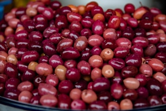 A pile of vibrant, glossy olives of varying shades of red and pink is displayed. The fruits are closely packed together, showcasing a rich, shiny surface that highlights their freshness.