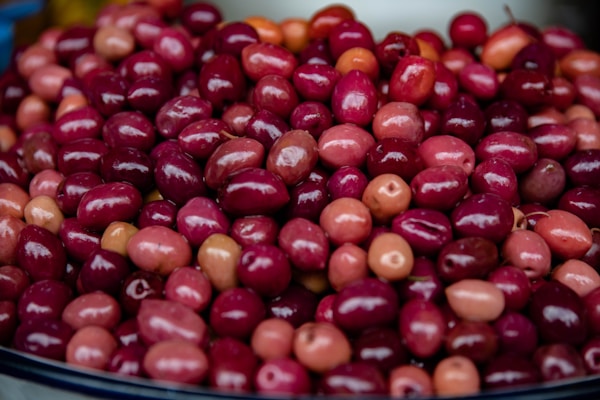 A pile of vibrant, glossy olives of varying shades of red and pink is displayed. The fruits are closely packed together, showcasing a rich, shiny surface that highlights their freshness.