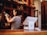 A person is examining a wine bottle in a warmly lit interior, possibly a wine shop or bar. Shelves filled with various bottles are visible in the background. The foreground features a modern point-of-sale terminal situated on a wooden countertop.