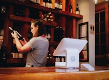 A person is examining a wine bottle in a warmly lit interior, possibly a wine shop or bar. Shelves filled with various bottles are visible in the background. The foreground features a modern point-of-sale terminal situated on a wooden countertop.