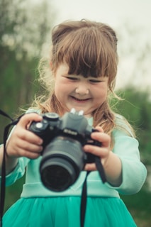 A young girl with light brown hair and bangs is smiling as she holds a large camera in her hands. She is wearing a teal dress, and appears to be outdoors, with a blurred green background suggesting a natural setting.