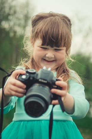 A young girl with light brown hair and bangs is smiling as she holds a large camera in her hands. She is wearing a teal dress, and appears to be outdoors, with a blurred green background suggesting a natural setting.