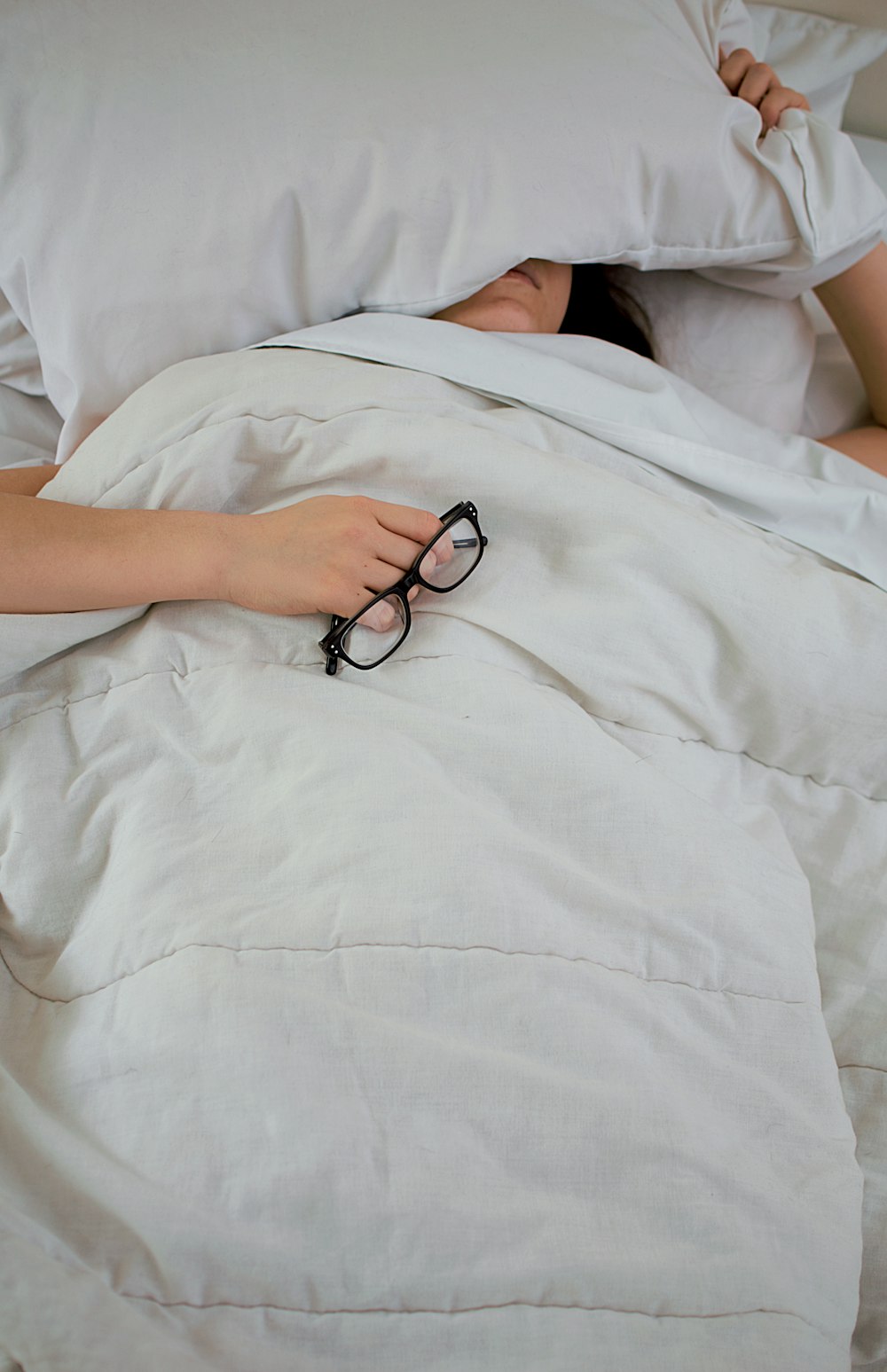 pessoa deitada na cama enquanto cobre o rosto com travesseiro e segura óculos