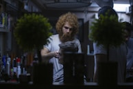 A barber with curly hair and a beard is giving a haircut to a seated customer in a dimly lit barbershop. The shop has various hair products on shelves and two potted plants in the foreground, which slightly obscure the view. Another person is partially visible in the background, possibly another customer or assistant.