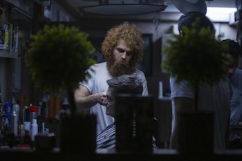 A barber with curly hair and a beard is giving a haircut to a seated customer in a dimly lit barbershop. The shop has various hair products on shelves and two potted plants in the foreground, which slightly obscure the view. Another person is partially visible in the background, possibly another customer or assistant.
