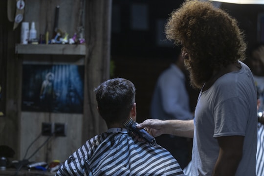 A barber is giving a haircut to a seated customer in a dimly lit barbershop. The customer wears a black and white striped cape. The room is decorated with a poster on the back wall and various items on a shelf. The barber has curly hair and a beard, wearing a white t-shirt.