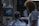 A barber is giving a haircut to a seated customer in a dimly lit barbershop. The customer wears a black and white striped cape. The room is decorated with a poster on the back wall and various items on a shelf. The barber has curly hair and a beard, wearing a white t-shirt.