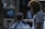 A barber is giving a haircut to a seated customer in a dimly lit barbershop. The customer wears a black and white striped cape. The room is decorated with a poster on the back wall and various items on a shelf. The barber has curly hair and a beard, wearing a white t-shirt.