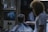 A barber is giving a haircut to a seated customer in a dimly lit barbershop. The customer wears a black and white striped cape. The room is decorated with a poster on the back wall and various items on a shelf. The barber has curly hair and a beard, wearing a white t-shirt.