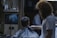 A barber is giving a haircut to a seated customer in a dimly lit barbershop. The customer wears a black and white striped cape. The room is decorated with a poster on the back wall and various items on a shelf. The barber has curly hair and a beard, wearing a white t-shirt.