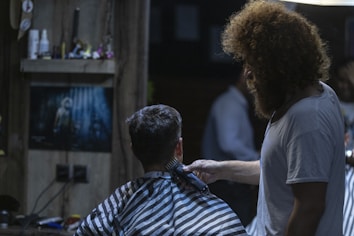 A barber is giving a haircut to a seated customer in a dimly lit barbershop. The customer wears a black and white striped cape. The room is decorated with a poster on the back wall and various items on a shelf. The barber has curly hair and a beard, wearing a white t-shirt.