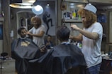 A man with curly hair and a beanie is giving a haircut to another man in a barbershop. Another customer is seated wearing a black cape, waiting for his turn. The barbershop is decorated and has various hair products and tools on the shelves. There is a bright light overhead and a mirror reflecting the scene.