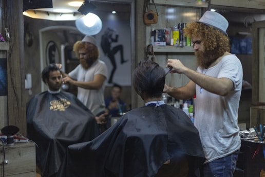 A man with curly hair and a beanie is giving a haircut to another man in a barbershop. Another customer is seated wearing a black cape, waiting for his turn. The barbershop is decorated and has various hair products and tools on the shelves. There is a bright light overhead and a mirror reflecting the scene.