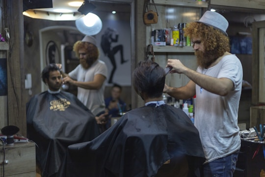 A man with curly hair and a beanie is giving a haircut to another man in a barbershop. Another customer is seated wearing a black cape, waiting for his turn. The barbershop is decorated and has various hair products and tools on the shelves. There is a bright light overhead and a mirror reflecting the scene.