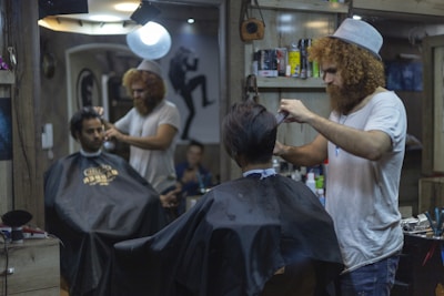 A man with curly hair and a beanie is giving a haircut to another man in a barbershop. Another customer is seated wearing a black cape, waiting for his turn. The barbershop is decorated and has various hair products and tools on the shelves. There is a bright light overhead and a mirror reflecting the scene.
