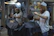 A man with curly hair and a beanie is giving a haircut to another man in a barbershop. Another customer is seated wearing a black cape, waiting for his turn. The barbershop is decorated and has various hair products and tools on the shelves. There is a bright light overhead and a mirror reflecting the scene.