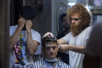 A man with curly hair and a beard is giving a haircut to another seated man in a barber's chair. The barber looks focused, while the client appears relaxed. Another man in a colorful shirt is standing nearby with his face partially covered by his hands. The setting is indoors with patterned walls and dim lighting.