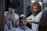 A man with curly hair and a beard is giving a haircut to another seated man in a barber's chair. The barber looks focused, while the client appears relaxed. Another man in a colorful shirt is standing nearby with his face partially covered by his hands. The setting is indoors with patterned walls and dim lighting.