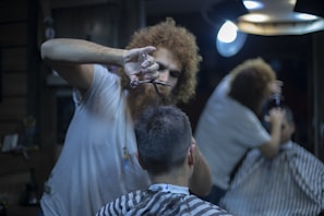 A barber, with curly hair, focuses on cutting a client's hair using scissors. The client is seated in a barber chair, wearing a striped cape, and facing away from the camera. The setting is dimly lit, with a mirror reflecting part of the scene and a round overhead lamp providing illumination.