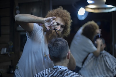 A barber, with curly hair, focuses on cutting a client's hair using scissors. The client is seated in a barber chair, wearing a striped cape, and facing away from the camera. The setting is dimly lit, with a mirror reflecting part of the scene and a round overhead lamp providing illumination.