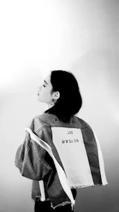 A black and white photograph featuring a person with short hair, wearing a denim jacket. The back of the jacket displays bold text reading 'NO FEAR' and other reversed text. The lighting creates a soft, artistic contrast.